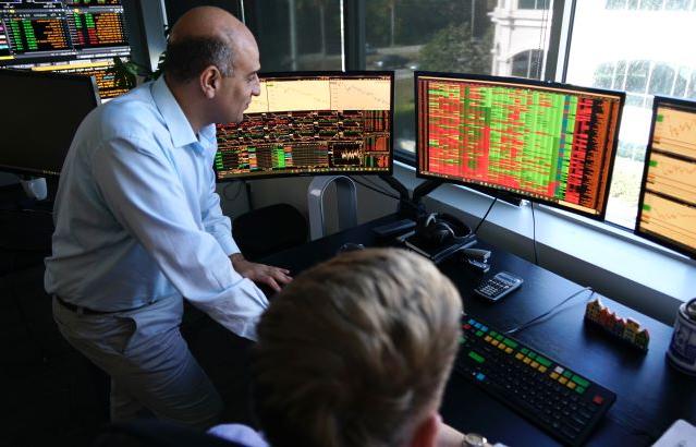 A nicely dressed male faculty member is working in the FinTech lab. He is standing beside a student and is in front of several dark monitors that have multicolored text on the screen.